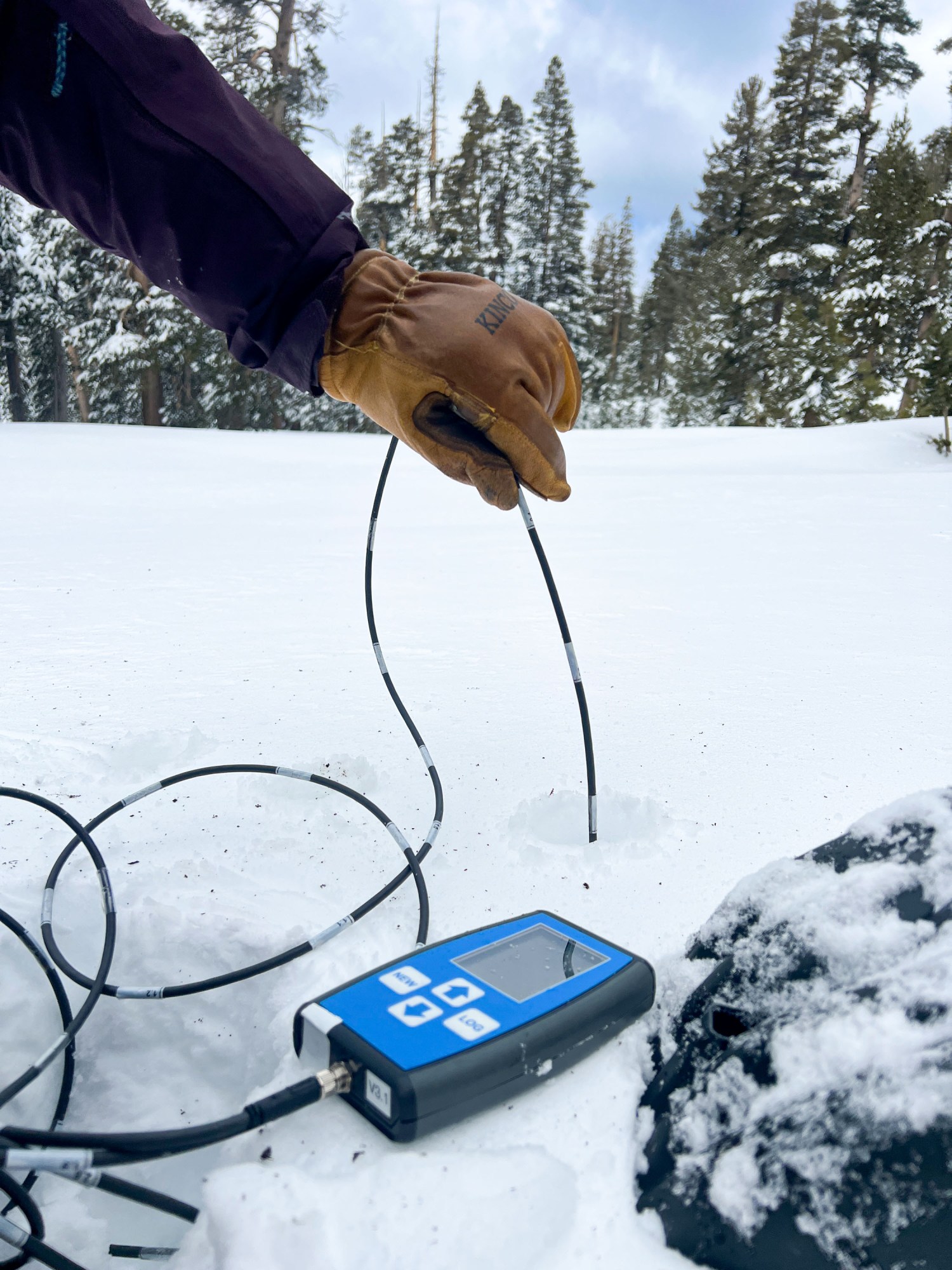 a gloved hand inserts a probe wire into a hole in the snow