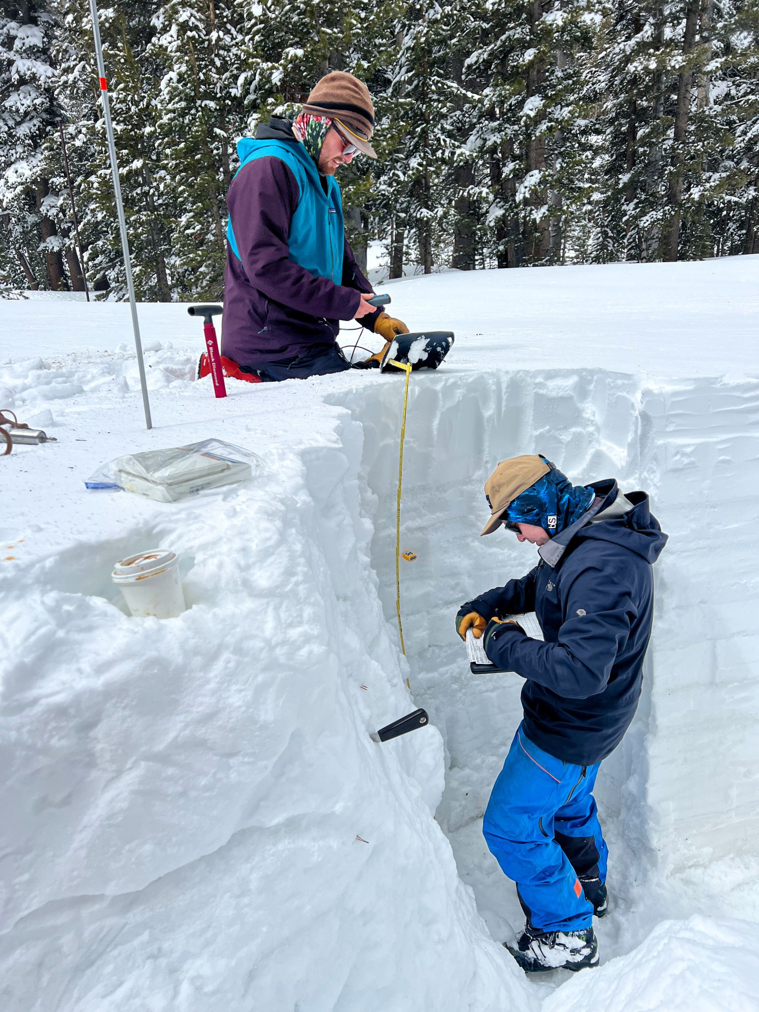 a researcher stands in a snowy trench taking notes, while a second researcher drops a yellow measure down from the surface level