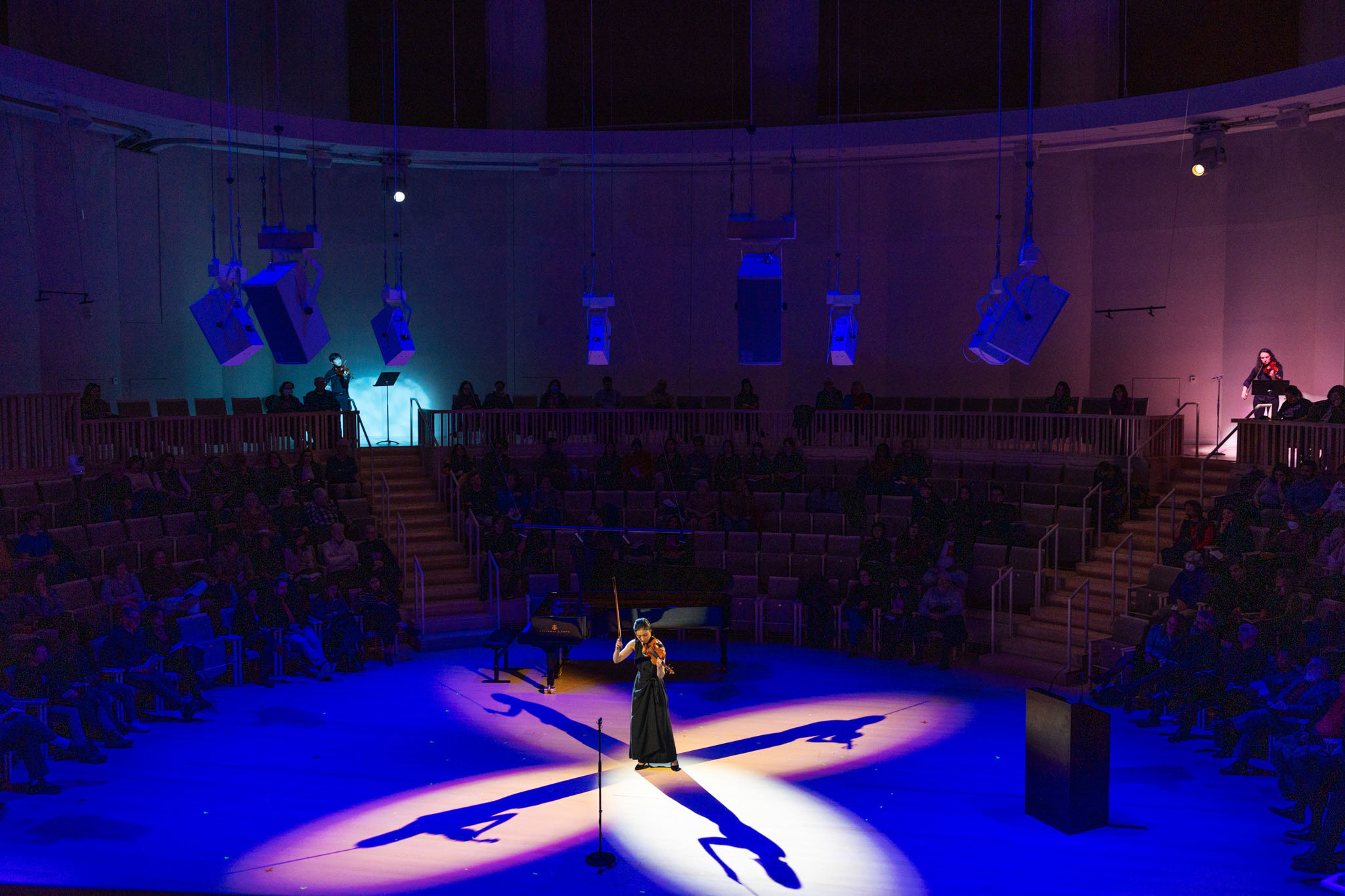 a woman holds her bow aloft as she plays the violin at the center of converging beams of the spotlights such that four shadows extend away from her at each 90 degree angle.