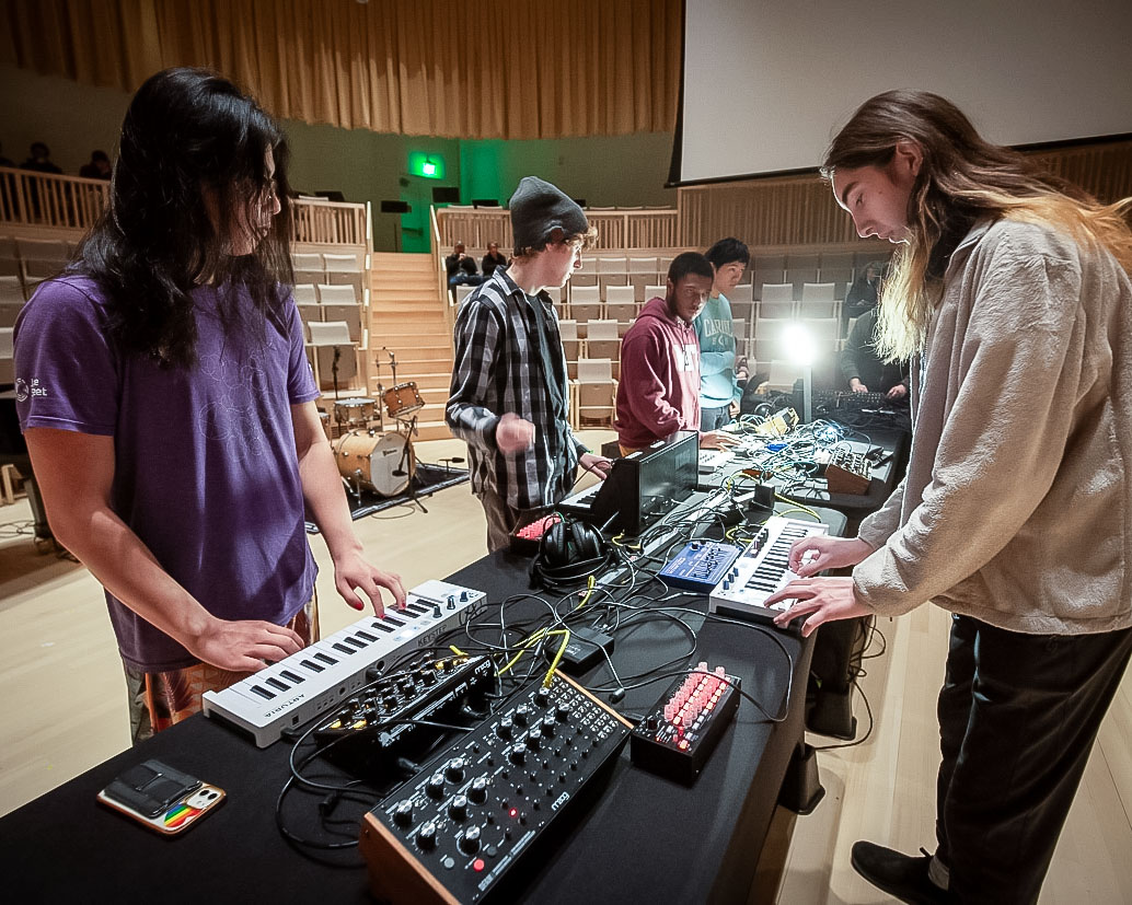Students on the performance floor stand at a long table with keyboards and other controllers