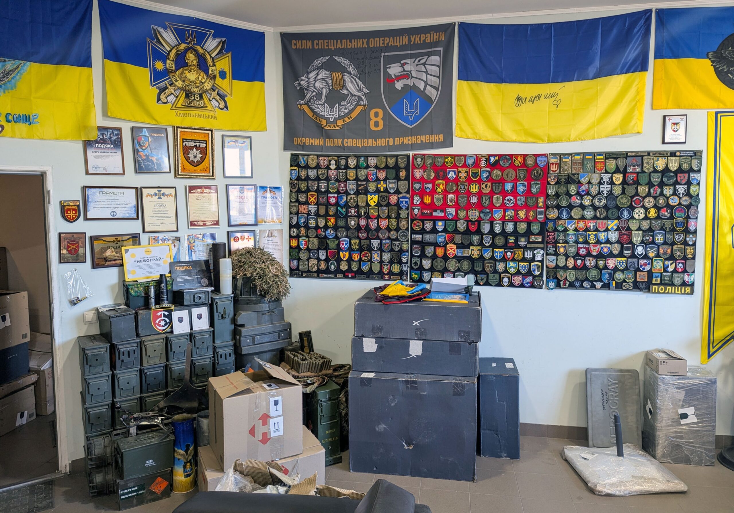A room with walls covered by a grid of patches and Ukrainian flags, and stacks of grey boxes on the floor