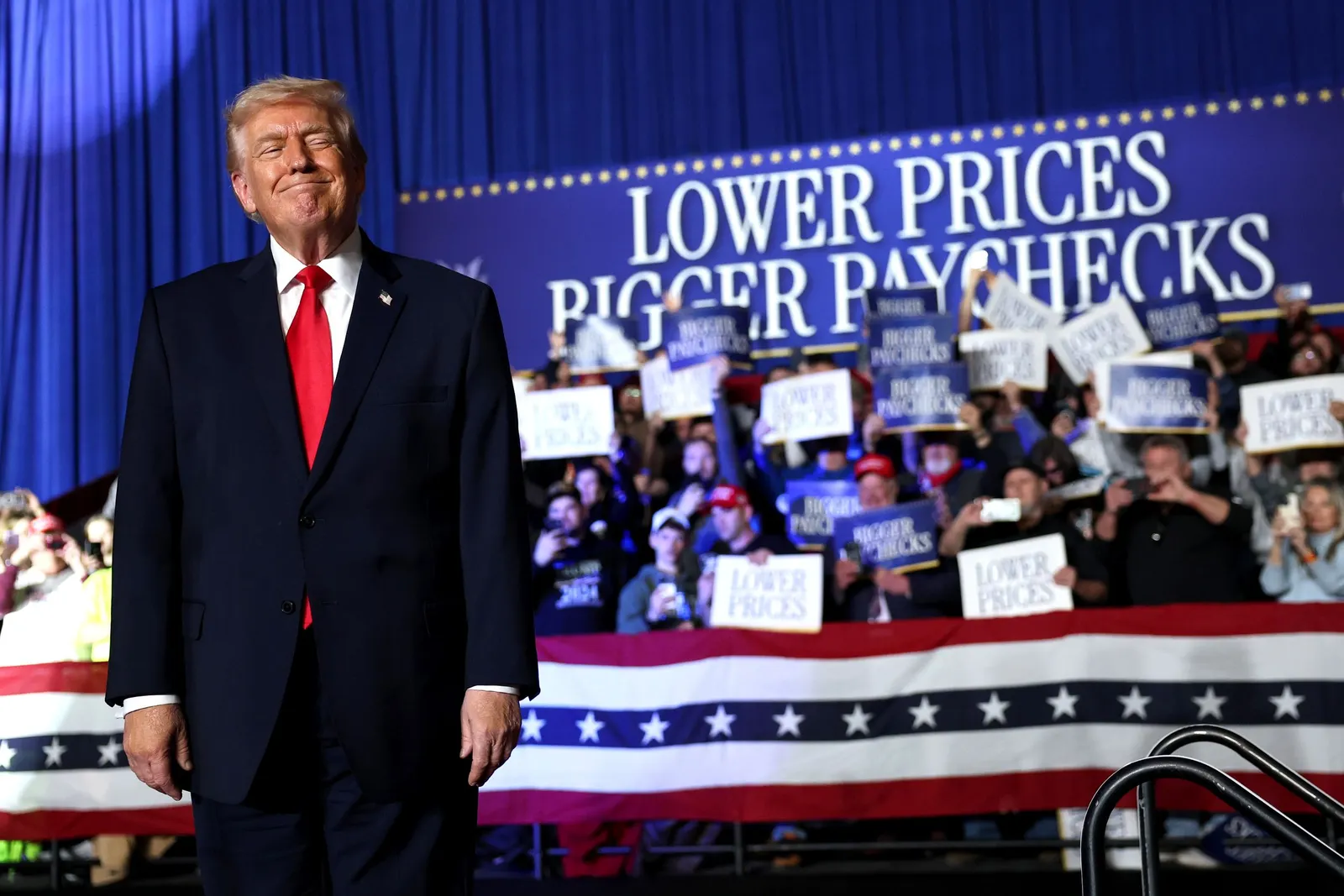 Donald Trump stands before a sign that says "lower prices, bigger paychecks"