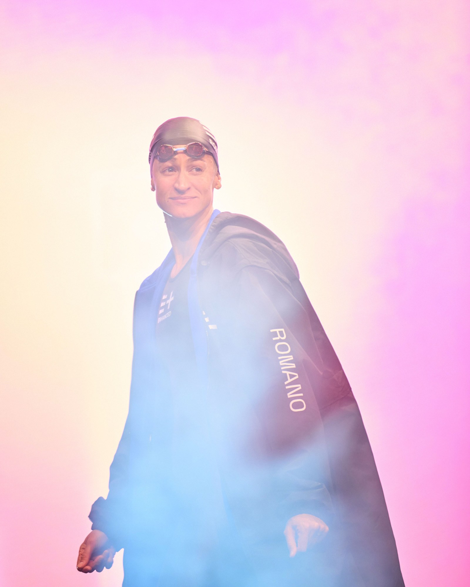 Megan Romano in cap and goggles with her dry robe stands backlit by pink event lighting and stage fog