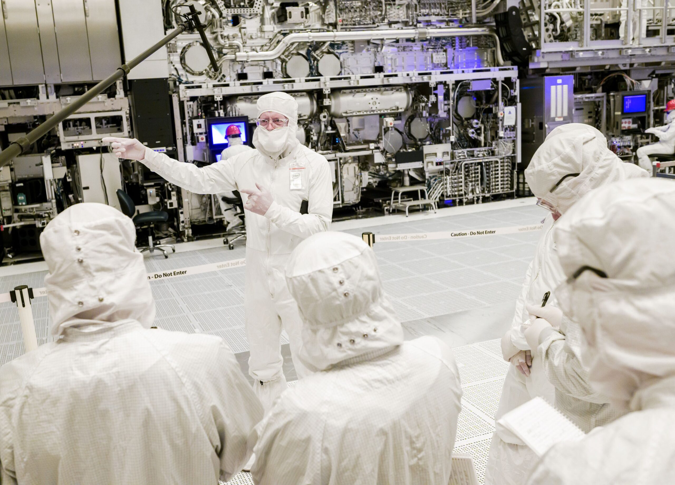over the shoulders of a crowd of workers in cleanroom suits listening to a central figure.